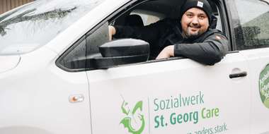 Eine Person sitzt am Steuer eines weißen Autos mit der Aufschrift „Sozialwerk St. Georg Care“.